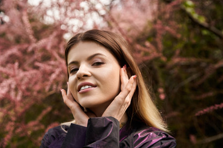 Beautiful woman smiles, hands on her face. Surrounded by pink blossoms.