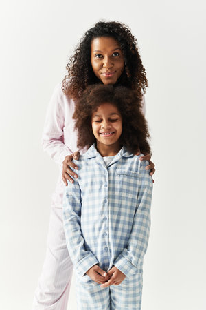 Black mother hugs daughter in matching PJs, grey background.