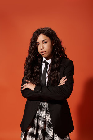 Confident young woman in a suit and plaid skirt poses against a bold orange backdrop.の写真素材