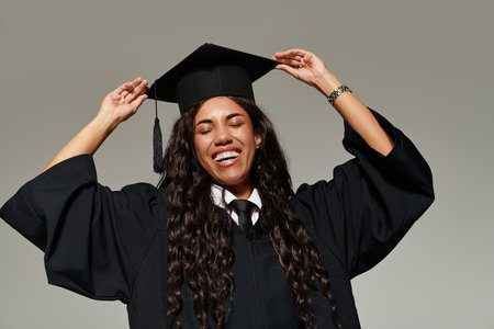 A young woman joyfully celebrates her graduation in a cap and gown, radiating happiness.の写真素材
