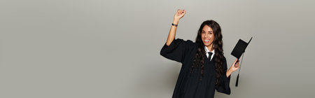A joyful young woman in a graduation cap and gown celebrates with raised arms.の写真素材