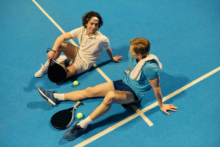 Two young sportsmen relax on the padel court, discussing strategy after a thrilling rally.の写真素材