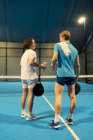 Two young sportsmen pause to chat and hydrate after an energetic padel match on a bright court.の写真素材