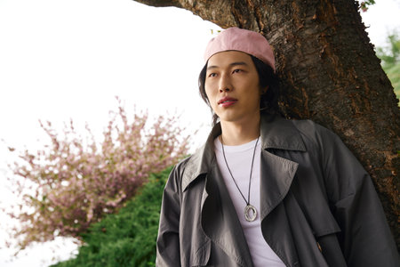 A young asian man relaxes in a vibrant park, showcasing a trendy grey outfit perfect for spring.