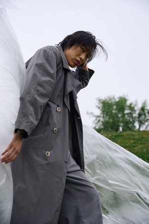 A stylish young Asian man stands confidently in a grey trench coat, covered in polyethyleneの写真素材