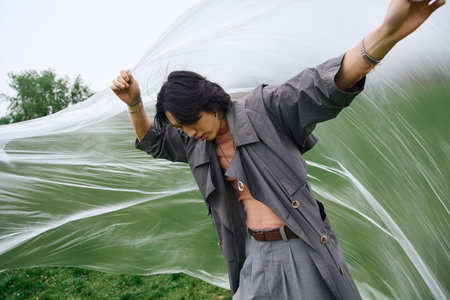 In a peaceful park, a young man in a grey trench coat expresses his unique fashion sense while holding polyethyleneの写真素材