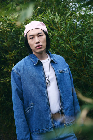 A stylish young Asian man confidently displays his denim outfit in a serene park environment.