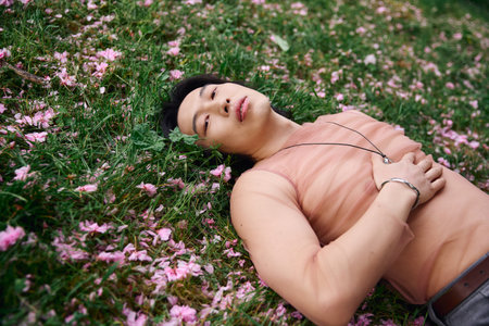A young Asian man relaxes on the grass, surrounded by blooming flowers in a stylish outfit.の写真素材