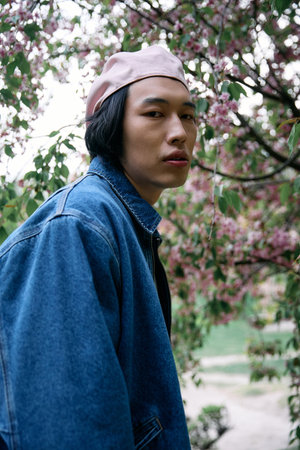 Young asian man in a fashionable denim outfit stands confidently in a lush park filled with flowers.
