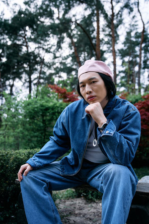 A young Asian man in a chic denim outfit showcases his style while relaxing in a park setting.の写真素材