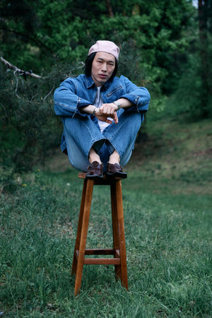 A young asian man sits comfortably on a stool, dressed in trendy denim attire, enjoying nature.