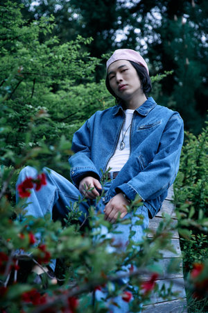 A young Asian man dressed in a fashionable denim outfit relaxes amidst vibrant greenery in a park.