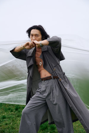 A young man wears a stylish grey trench coat and poses confidently near polyethylene in a peaceful...