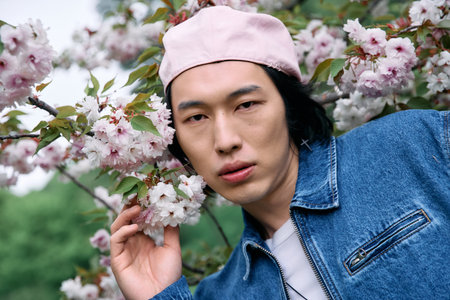 Young asian man poses confidently in denim outfit among vibrant cherry blossoms in spring.