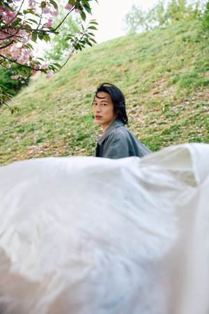 A young Asian man in a stylish grey trench coat posing with polyethylene in a green park.の写真素材