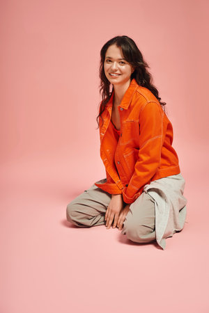 Young woman sits gracefully in a trendy outfit, radiating confidence against a soft pink backdrop.