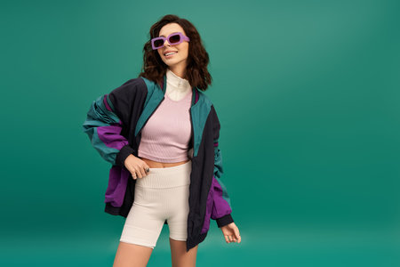 A stylish young woman confidently poses in a sporty outfit against a turquoise backdrop.
