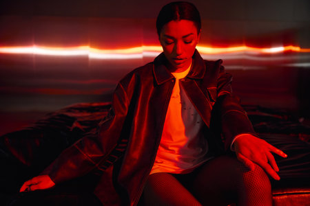 Chic young woman dressed in fashionable attire sits gracefully in a striking red lit environment.の写真素材