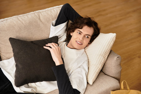 A young man lounges on his sofa, holding a pillow and wearing casual home attire.