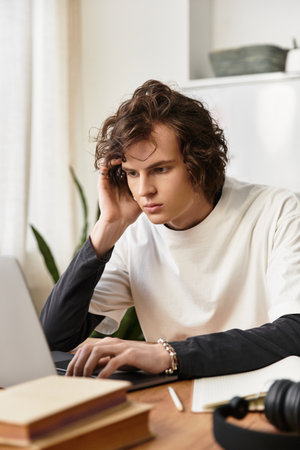 A young man in casual clothes focuses on his laptop at a cozy desk.