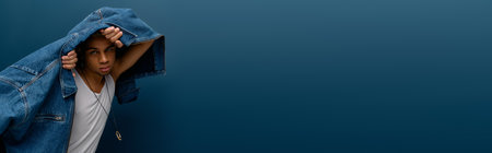 Stylish young African American man poses confidently in a denim jacket on a solid blue background, bannerの写真素材