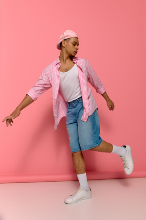 A young Black man in a checkered shirt and beret poses against a bright pink background.