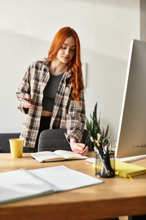 A young woman with long red hair writes in her notebook at her cozy home office.の写真素材