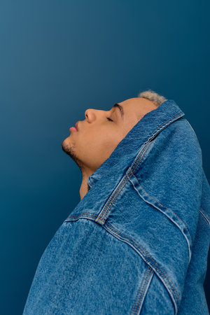A young Black man shows off his dyed hair and style in a denim jacket by a blue wall.の写真素材