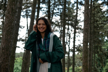 A young woman with curly hair smiles while wearing a fashionable raincoat in a vibrant forest.の写真素材
