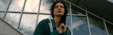 Curly-haired woman poses confidently in a trendy raincoat amidst a modern outdoor setting, banner