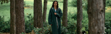 A young woman with curly hair stands gracefully in a vibrant forest wearing a fashionable raincoat, bannerの写真素材