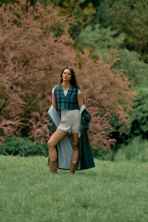 Confident young woman with curly hair poses in a stylish raincoat surrounded by vibrant nature.