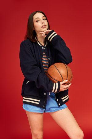 A stylish young woman poses with a basketball in trendy attire against a bold backdrop.