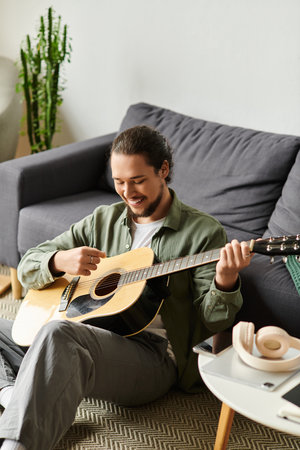 Handsome young man sits on the floor, strumming his guitar, lost in music at home.