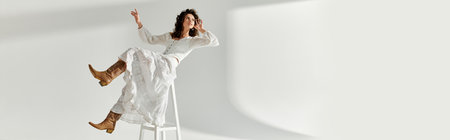 Stylish young woman poses confidently on a stool dressed in a flowy white outfit and boots.