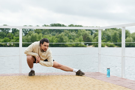 A young athletic man performs a stretching exercise outdoors near a tranquil body of water.