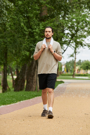 A bearded young man walks along a path in a green park, towel around his neck, exercising.