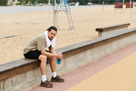 A young man rests on a bench, enjoying a drink after exercising outdoors near the beach.の写真素材