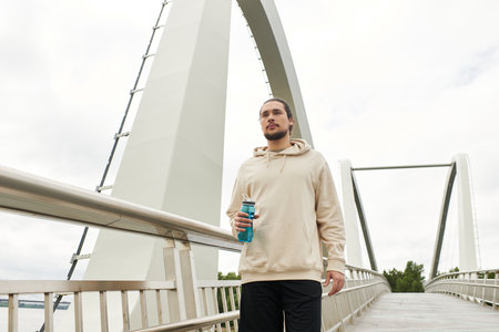 A young man in a hoodie works out on a beautiful bridge, staying hydrated.