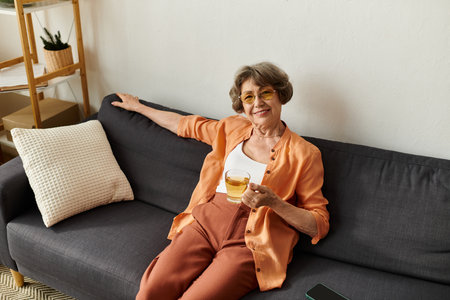 A senior woman in cozy clothes lounges comfortably on her sofa, sipping a warm drink and smiling.の写真素材