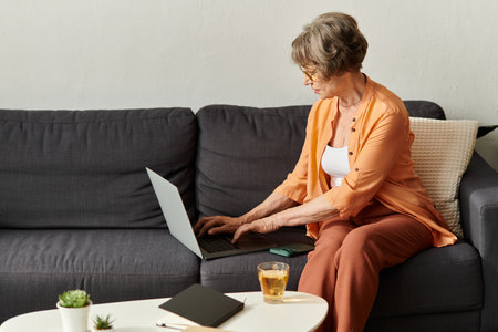 A senior woman in comfy attire sits on a couch, using her laptop, enjoying a warm drink.