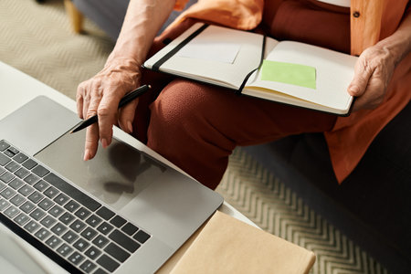 Mature woman in cozy attire focused on her laptop while taking notes in her notebook at homeの写真素材
