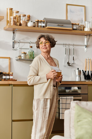 A mature woman in comfortable attire relaxes with a warm drink in her inviting kitchen space.の写真素材