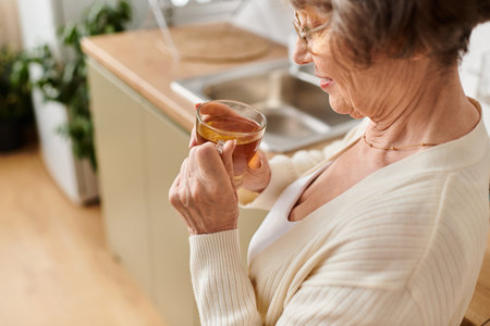 A mature woman in cozy attire enjoys her warm drink in the comfort of her kitchen.の写真素材