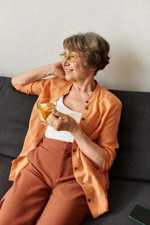 A senior woman in cozy attire enjoys a warm drink on the sofa with a smile.の写真素材