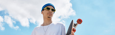 A young man stands confidently in a park holding his skateboard, basking in the sunlight.