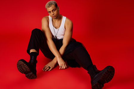 A young Black man in a tank top and black pants poses stylishly in a red studio.