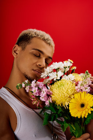 A young man in a tank top embraces colorful flowers against a striking red background.の写真素材
