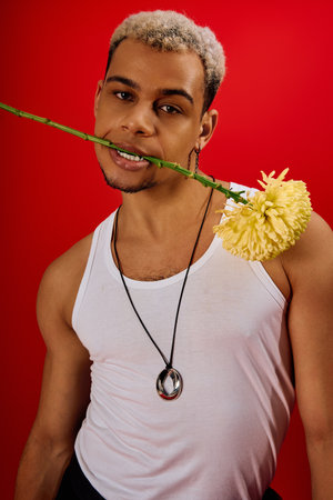 A young African American man poses confidently in a red studio with a flower in his mouth.
