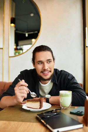 A young man with a beard enjoung matcha and cake in a cozy cafe.